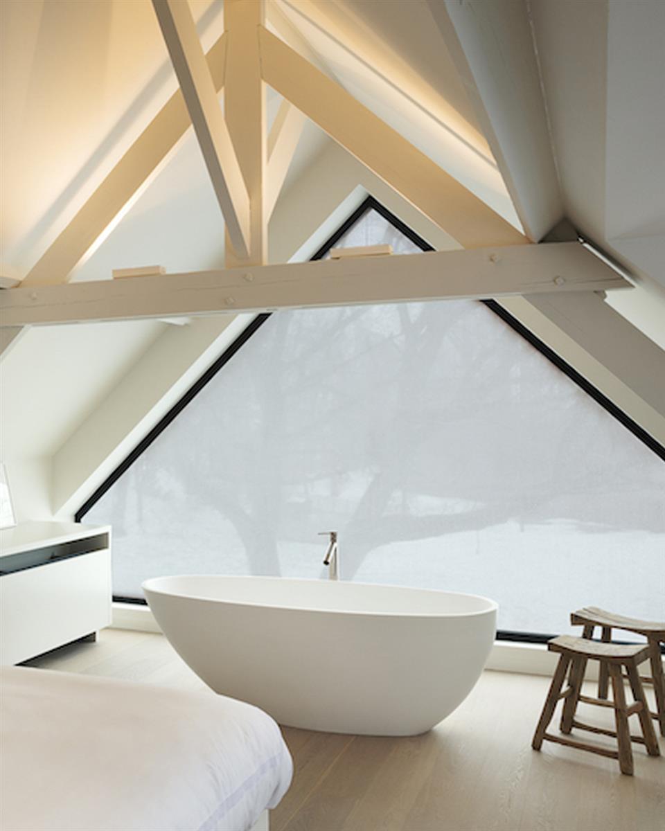 Baderom med trekantet vindu med Squid vindutekstil, frittstående badekar, to krakker og takbjelker. Foto.
